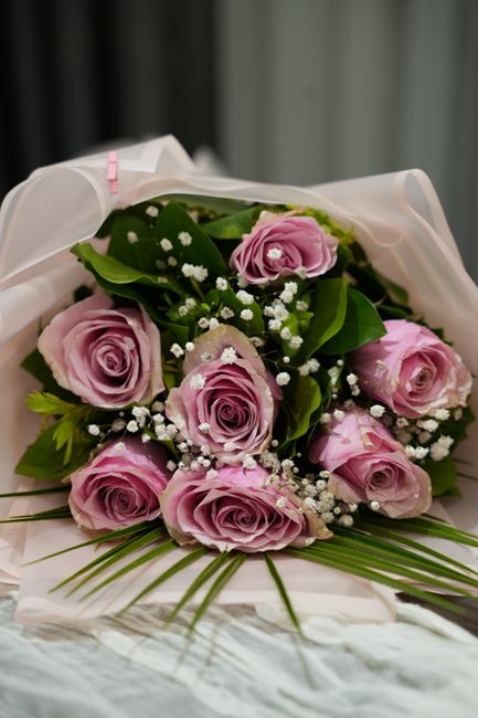 A bouquet of fresh pink roses with delicate, tightly curled petals, accented by small white baby's breath flowers and lush green foliage, arranged in a classic floral style. The roses are partially wr