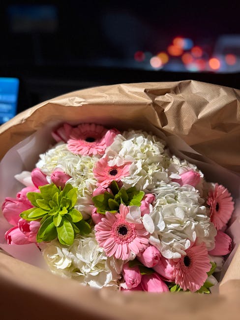 A bouquet of fresh flowers held inside a brown paper wrapping, prominently featuring white hydrangeas, pink gerbera daisies, and small green foliage accents. The flowers are arranged in a rounded, lus