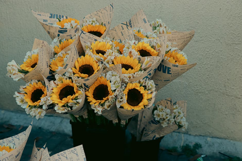 A bouquet of fresh sunflowers with bright yellow petals and dark centers, surrounded by small white daisies. The flowers are wrapped in brown paper with printed text and arranged in a black vase, plac