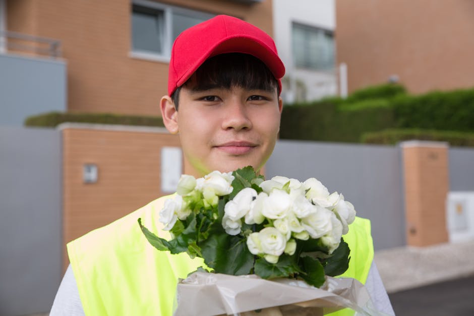 A young male flower delivery courier wearing a red cap and a high-visibility yellow vest is holding a bouquet of white roses wrapped in white paper. The bouquet features fresh, tightly closed blossoms