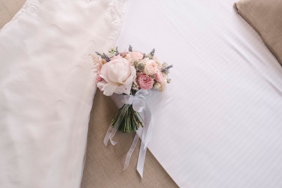 A delicate bouquet of fresh flowers resting on a white bedspread, featuring soft pink and white roses, peonies, and small lavender sprigs, all arranged in a white paper wrap tied with a sheer white ri
