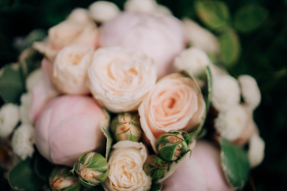 A close-up of a floral bouquet featuring soft pink and cream-colored roses with ruffled petals and green stems, surrounded by small white filler flowers and dark green leaves. The flowers are arranged