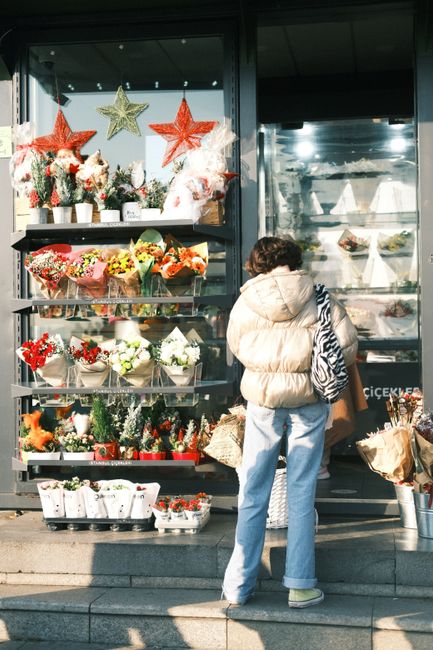 A woman wearing a beige puffer jacket, light blue jeans, and yellow sneakers stands outside a flower shop, viewing an array of floral arrangements displayed on a glass shelf. The flowers include red, 